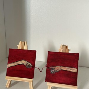 Puede incluir: Dos pequeñas pinturas cuadradas sobre caballetes de madera. Cada pintura presenta una mano extendida sobre un fondo rojo, conectadas por un hilo. La obra de arte es una versión en miniatura de la famosa pintura "La Creación de Adán".