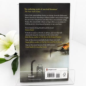 May include: A book cover for "Man's Search for Meaning" by Viktor Frankl, displayed in a clear acrylic stand. The cover features text from The New York Times and other authors. The book's cover has a dark background with a graphic of a bird and a wheelchair.