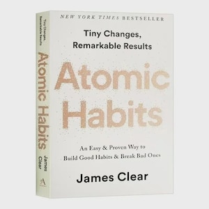 May include: A hardcover book titled "Atomic Habits" by James Clear. The cover is white with the title in large, pixelated, copper-colored letters. The book is a New York Times bestseller, with the subtitle "An Easy & Proven Way to Build Good Habits & Break Bad Ones."