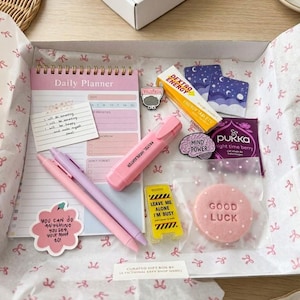 May include: A gift box with a pink and white theme, containing a daily planner, pens, highlighter, stickers, energy tablets, tea, and a cookie with "Good Luck" printed on it. The box is lined with tissue paper and bows.