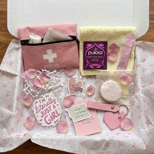 Pode incluir: Uma caixa de presente cheia de itens rosa, incluindo um kit de primeiros socorros, uma toalha pequena, chá, um biscoito, adesivos e um chaveiro em forma de coração. A caixa é forrada com papel estampado e salpicada com pétalas de rosa rosa.