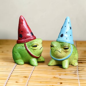 May include: Two quirky green frog ornaments with grumpy faces. One frog wears a red hat with black floral details, the other a blue hat with white star and floral details. Both have a matching collar. The ornaments are on a bamboo mat.