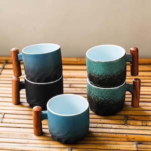 May include: A set of three ceramic mugs with a speckled, cream-colored glaze. Each mug features a different hand-painted floral design in shades of blue, green, and yellow. The mugs have a simple, rounded shape and a comfortable handle.