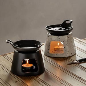 Pode incluir: Dois aquecedores de óleo de cerâmica. Um é preto, o outro branco com salpicos pretos. Cada um tem um pequeno prato em cima e uma abertura retangular para segurar uma vela de chá acesa. A chama da vela é visível.