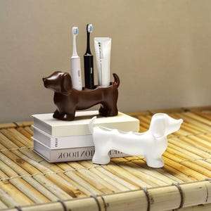 Puede incluir: Un soporte para cepillos de dientes con forma de perro salchicha de cerámica marrón con un cepillo de dientes blanco y negro, y un tubo de pasta de dientes. El soporte está encima de una pila de libros. Un soporte blanco con forma de perro salchicha está en primer plano.