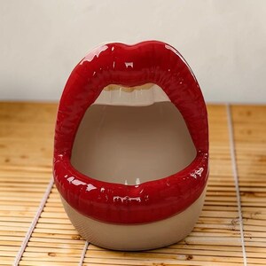 Könnte beinhalten: Eine dekorative Keramikschale in Form roter Lippen. Die Schale hat eine glänzende Oberfläche und einen neutralen Innenraum. Der Boden der Schale ist neutralfarben, und die Lippen sind leuchtend rot.