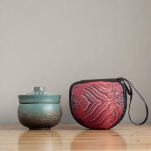 Könnte beinhalten: Eine türkisfarbene Keramik-Teetasse mit Deckel und eine rot gemusterte Tragetasche mit schwarzem Reißverschluss und Griff. Die Teetasse hat eine strukturierte Oberfläche und einen kleinen Deckel.