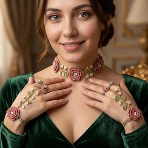 May include: A crochet floral jewelry set featuring a choker necklace, bracelet, ring, and hand chain. The set is made of pink flowers, green leaves, and white accents, with a sparkling center. The woman is wearing a green velvet dress.