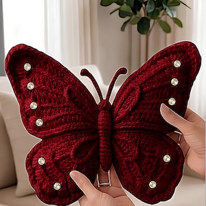 Könnte beinhalten: Eine große, weinrote Schmetterlingsdekoration. Der Schmetterling besteht aus einem Strickmaterial und ist mit funkelnden, runden Verzierungen versehen. Die Flügel sind mit strukturierten Mustern versehen, und die Fühler sind gebogen.