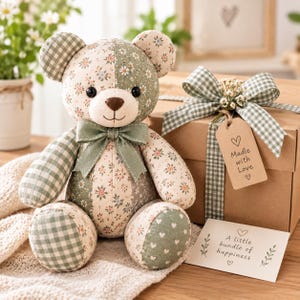 May include: A patchwork teddy bear made of sage green and cream fabric with floral and gingham patterns, featuring a matching green ribbon bow at the neck. Beside it sits a brown gift box with a green gingham ribbon and a tag reading Made with Love.