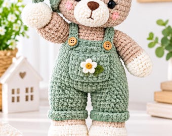 May include: A crocheted teddy bear wearing a sage green hat with a white flower, and sage green overalls with two brown buttons and a white flower on the chest pocket. The bear has light brown fur, white paws, and pink cheeks. Text reads 70% descount first 40 buyer only.