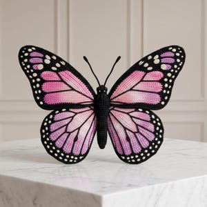 Może przedstawiać: Ręcznie wykonany motyl z czarnym korpusem i skrzydłami. Skrzydła mają gradient różu i fioletu, obrysowane na czarno z białymi kropkami. Motyl jest prezentowany na białej marmurowej powierzchni.