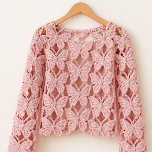 May include: A light pink crochet top featuring a butterfly design. The long-sleeved top is made of a delicate, open-weave fabric. Each butterfly has a small, silver-colored embellishment in the center. The top is hanging on a wooden hanger.