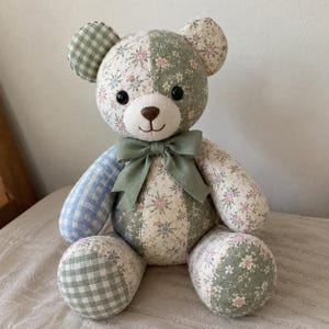 May include: A stuffed teddy bear crafted from various fabric patches featuring floral patterns, green and white gingham, and blue and white plaid. The bear has black button eyes, a brown stitched nose, and a sage green ribbon tied around its neck.