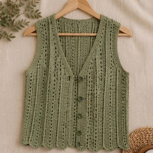 May include: A sage green sleeveless crochet vest featuring a V-neckline, a vertical ribbed pattern with openwork detailing, and a scalloped hem. The front of the garment is secured with five matching green buttons arranged in a single vertical row.