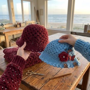 May include: Two crochet hats are displayed. One is burgundy with white embellishments, and the other is light blue with red flowers and butterflies. The hats are on a wooden table with a pair of scissors and a crochet hook.