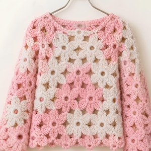 May include: A pink and white crochet sweater with a floral pattern. The long-sleeved sweater is made of yarn and features a repeating flower design. The sweater is hanging on a silver hanger.
