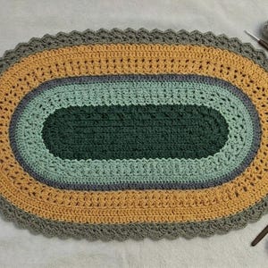May include: A handmade, oval crochet rug in shades of grey, yellow, green, and blue. The rug has a scalloped edge and a layered design, with a dark green centre and a yellow outer layer. Two crochet hooks and a ball of yarn are next to the rug.