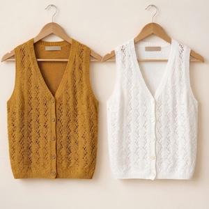 May include: Two sleeveless V-neck sweater vests hanging on wooden hangers. One vest is mustard yellow and the other is white. Both feature a vertical openwork knit pattern, a button-down front with matching buttons, and a ribbed hem at the waist.