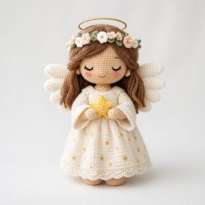Puede incluir: Muñeca de ángel de ganchillo con cabello castaño, ojos cerrados y una corona floral. La muñeca viste un vestido color crema con estrellas amarillas y sostiene una estrella amarilla. Tiene alas blancas y un halo dorado.