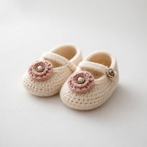 May include: A pair of cream-colored crochet baby shoes. Each shoe features a pink flower with a pearl center and a strap with a button closure. The shoes are designed with a Mary Jane style.