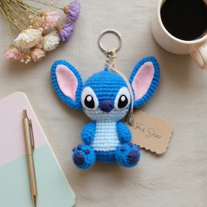 May include: A blue crochet Stitch keychain with large ears, pink inner ears, and a white belly. The keychain is attached to a metal ring. A small tag that says "For You" is next to the keychain.