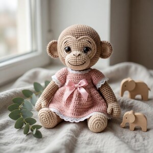Könnte beinhalten: Gehäkelte Affenpuppe mit hellbraunem Körper und rosa Kleid mit weißer Spitzenborte und rosa Schleife. Zwei kleine Holz-Elefantenfiguren liegen daneben. Das Ganze liegt auf einer grauen Oberfläche.