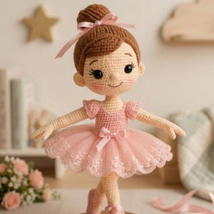 Könnte beinhalten: Eine handgefertigte Häkel-Ballerina-Puppe. Die Puppe hat braunes Haar in einem Dutt mit rosa Schleife, rosige Wangen und schwarze Augen. Sie trägt ein rosa Kleid mit Tüllrock und Ballettschuhen. Die Puppe steht mit ausgestreckten Armen.