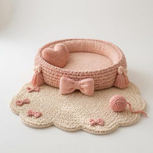 Op de afbeelding: Een roze gehaakt dierenbed met een hartvormig kussen, strik en kwastjes. Het bed staat op een crèmekleurige, geschulpte mat met kleine roze strikjes en een bolletje wol. Een gezellig en stijlvol ontwerp voor huisdieren.