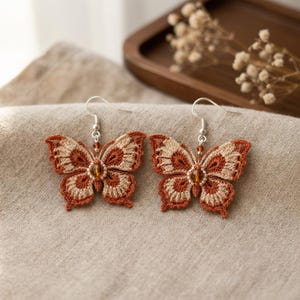 Puede incluir: Un par de pendientes de mariposa hechos a mano. Los pendientes presentan un diseño de ganchillo en tonos marrones y beige, con una pequeña cuenta en el centro. Los pendientes tienen ganchos plateados.