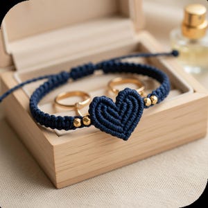 May include: A navy blue macrame bracelet with a heart-shaped design and gold-colored beads. The bracelet is displayed in a wooden box with two gold rings. A perfume bottle is visible in the background.