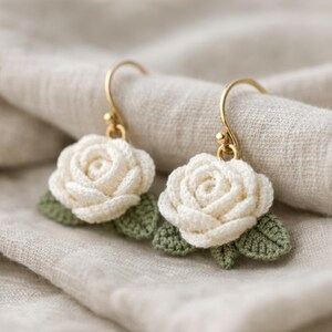Könnte beinhalten: Ein Paar handgefertigte Häkelohrringe mit elfenbeinfarbenen Rosenblüten mit grünen Blättern. Die Ohrringe haben goldfarbene Ohrhaken. Das florale Design ist vor einem neutralen Stoffhintergrund platziert.
