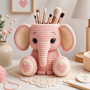 May include: A pink crocheted elephant-shaped container holding makeup brushes. The elephant has large ears, a trunk, and round feet with heart-shaped designs. The container is sitting on a white lace doily.