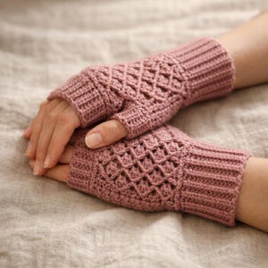 May include: A pair of mauve-colored fingerless gloves, crocheted with a diamond pattern on the hand and a ribbed cuff. The gloves are displayed on a neutral-toned fabric, showcasing the handmade texture and design.