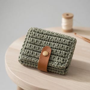 Könnte beinhalten: Eine salbeigrüne Häkeltasche mit einem braunen Lederriemen und einem goldfarbenen Knopfverschluss. Die Geldbörse ist rechteckig und hat ein strukturiertes Aussehen. Eine Garnrolle und eine Häkelnadel befinden sich im Hintergrund.