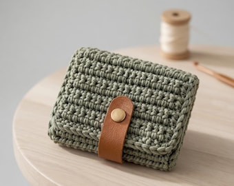 May include: A sage green crochet wallet with a brown leather strap and a gold-colored button closure. The wallet is rectangular and has a textured appearance. A spool of thread and a crochet hook are in the background.