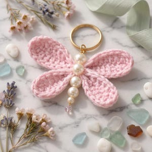 May include: A handmade pink crochet dragonfly keychain with pearl accents and a gold-colored ring. The dragonfly's wings are a soft pink, and the body is adorned with several faux pearls. Decorative elements include flowers and sea glass.