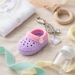 Può includere: Scarpina da neonato all'uncinetto lilla e rosa, con un bottone bianco. L'immagine include un biberon, un anello per la dentizione in legno, un body per neonato e un portachiavi con un ciondolo a forma di cuore, a tema baby.