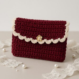May include: A burgundy crochet clutch with a scalloped, cream-colored trim. The flap is secured with a gold-tone flower clasp. The handmade bag is a stylish accessory for carrying essentials. Pearls and lace are in the background.