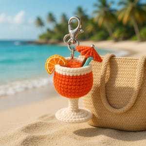 May include: A handmade orange cocktail keychain with a silver clasp, orange slice, and small orange umbrella. The drink sits on a white base. A woven beige tote bag is on a sandy beach with palm trees and ocean in the background.