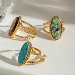 May include: Three gold-colored rings with oval-shaped gemstones. One ring features a brown stone, another a green stone, and the third a turquoise stone. The rings have a textured band design.