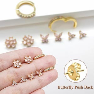 May include: A collection of gold-colored earrings with various designs, including flowers, butterflies, and small hoops. The earrings are adorned with small, clear and pink gemstones. The image also shows a close-up of a butterfly push back earring clasp.
