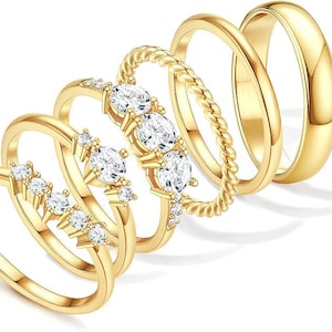 May include: A collection of six gold-colored rings. Some rings feature clear, sparkling gemstones, while others have a smooth, polished finish or a twisted rope design. The rings are arranged in a slightly overlapping pattern.