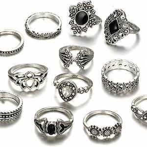 May include: A collection of silver-toned rings with various designs, including floral, geometric, and gemstone accents. The rings are arranged on a white surface, showcasing their intricate details and vintage style.