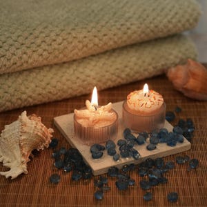 May include: Two lit candles, one starfish-shaped and the other floral, sit on a small square stone. Blue glass beads surround the candles. A seashell and folded towels are in the background, creating a spa-like ambiance.