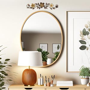 May include: An oval gold-framed mirror hangs above a wooden console table. A decorative floral metal piece is above the mirror. A lamp with a brown ribbed base and white shade sits on the table, along with art prints and potted plants. The room has a neutral color palette.