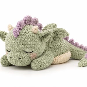 Op de afbeelding: Een gehaakte drakenknuffel in een slapende houding. De draak is voornamelijk saliegroen met een crèmekleurige buik, kleine crèmekleurige hoorns en een lavendelkleurige stekelige kam langs de rug en staart. Het heeft gesloten ogen en roze wangen.