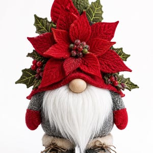 Op de afbeelding: Een decoratief kabouterfiguur met een rode vilten muts versierd met een grote rode kerstster, groene hulstbladeren en rode bessen. De kabouter heeft een lange witte baard van imitatiebont, een houten neus en grijze vilten laarzen met veters.
