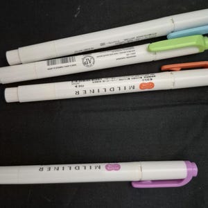 May include: Four Zebra Mildliner double-ended markers with white barrels. The markers feature caps and accents in light blue, light green, orange, and purple. Each marker displays the text MILDLINER and includes a barcode and product information.