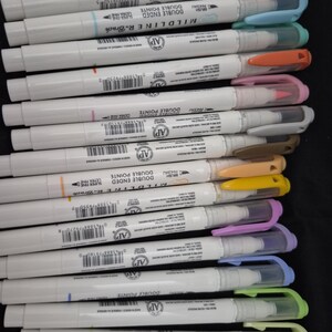 May include: A collection of Zebra Mildliner double-ended markers featuring white barrels with colored caps and clips. Each marker includes a brush tip and an ultra-fine tip. The assortment displays a variety of ink colors including pink, blue, orange, yellow, green, and purple, with visible barcodes and product text printed on the white plastic bodies.